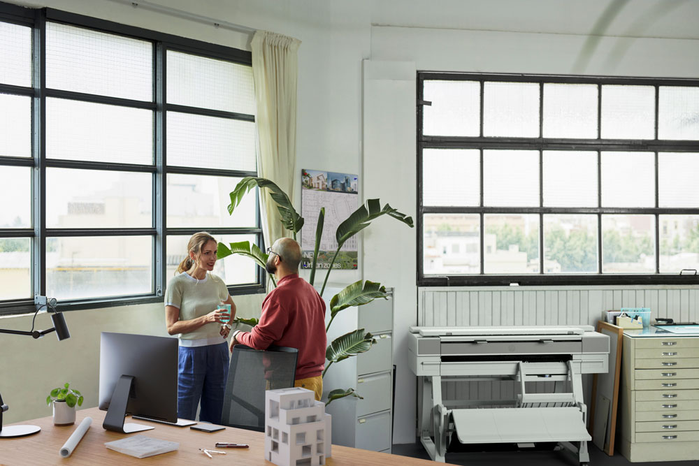 HP DesignJet XT950 printer placed in a bright architectural office while two colleagues discuss plans near a desk filled with design tools.